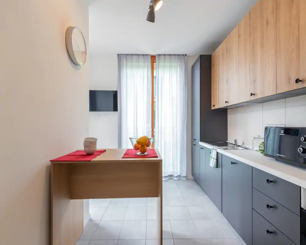 Modern compact kitchen with matte grey lower cabinets and warm wood upper cabinets, a white countertop, microwave and a small breakfast bar with fruit bowl. Natural light filters through sheer curtains over a balcony/door, creating a bright, functional space.