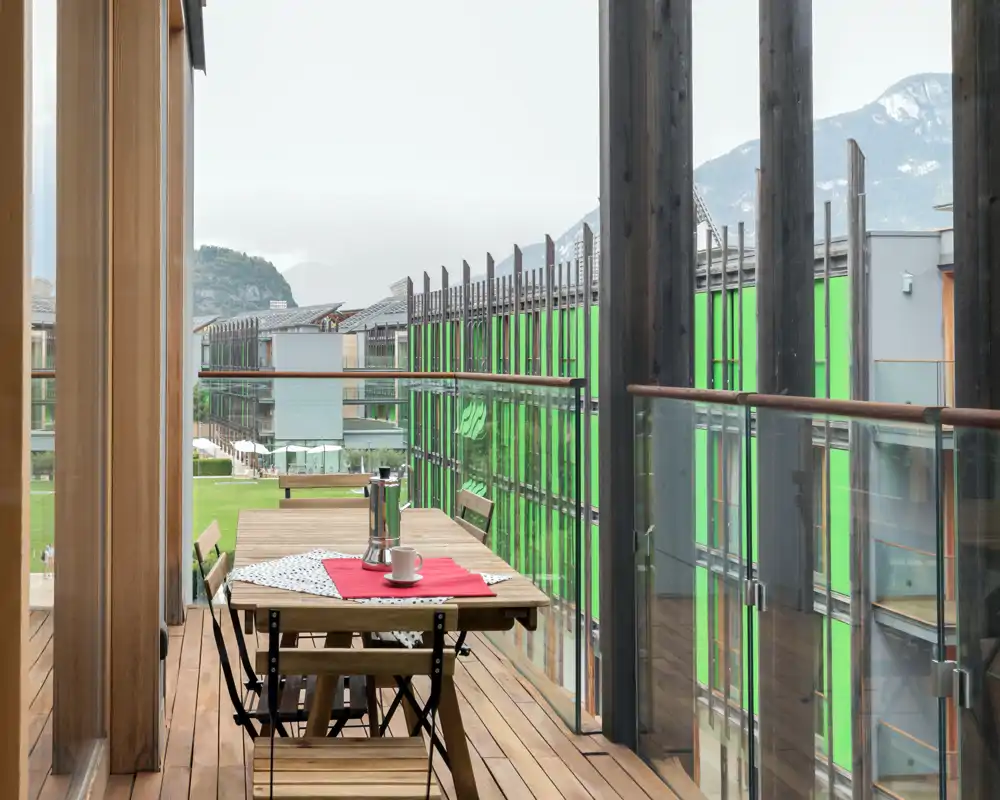 Bright wooden balcony with a small dining table, two foldable chairs, and glass railing offering views of neighboring modern buildings and distant mountains.