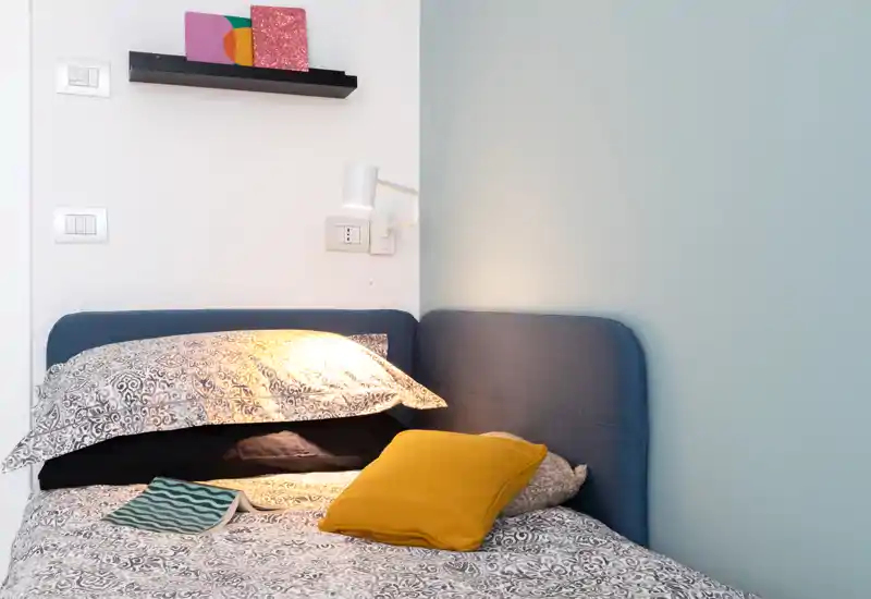 Bright, cozy bedroom corner with patterned bedding, layered pillows and a bedside reading light creating a warm, inviting atmosphere.