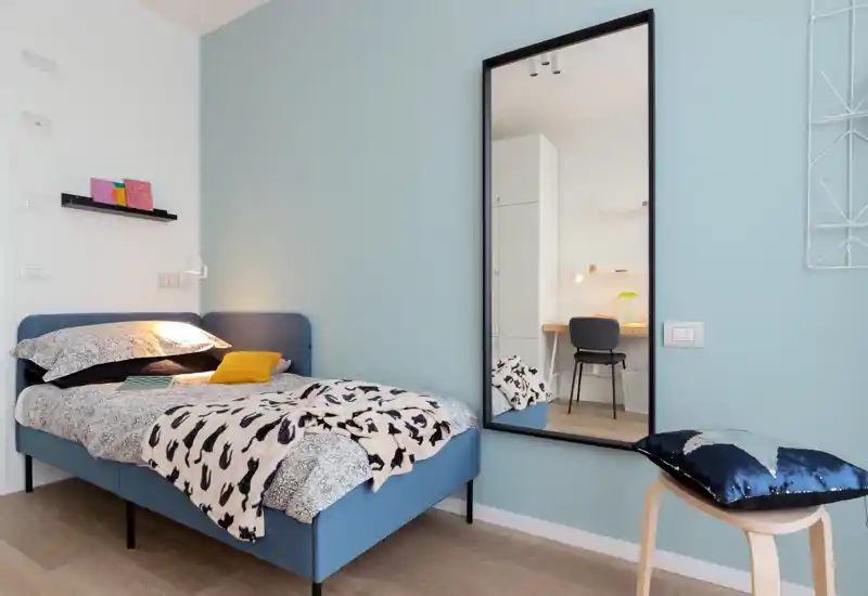 Bright, modern single bedroom with a blue upholstered bed, patterned bedding and a large wall mirror reflecting a neat workspace. Soft textiles and warm lighting create a cozy, well-styled space ideal for rental photos.