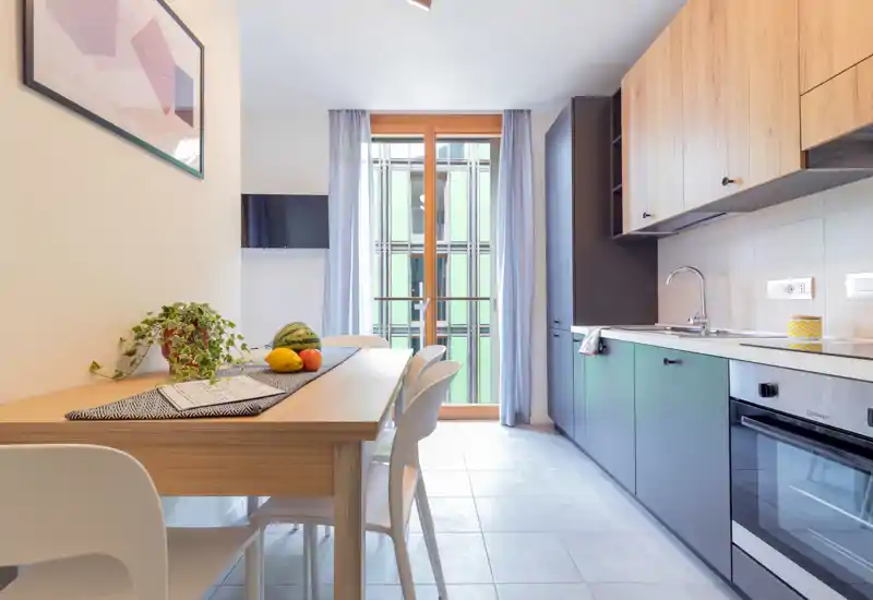 Bright, modern open-plan kitchen with integrated cabinets, a built-in oven and a wooden dining table set in front of a large window/door letting in natural light.