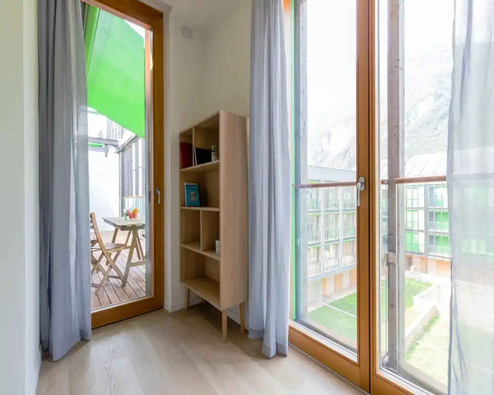 Bright corner living area with large floor-to-ceiling windows and a small wooden shelving unit; access to a balcony with a folding table and chairs and abundant natural light.