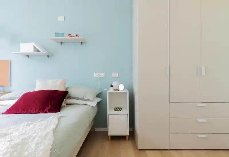 Bright, modern bedroom with a neatly made bed, soft pastel walls and a compact bedside cabinet next to a large wardrobe — clean, minimalist styling ideal for rental photos.