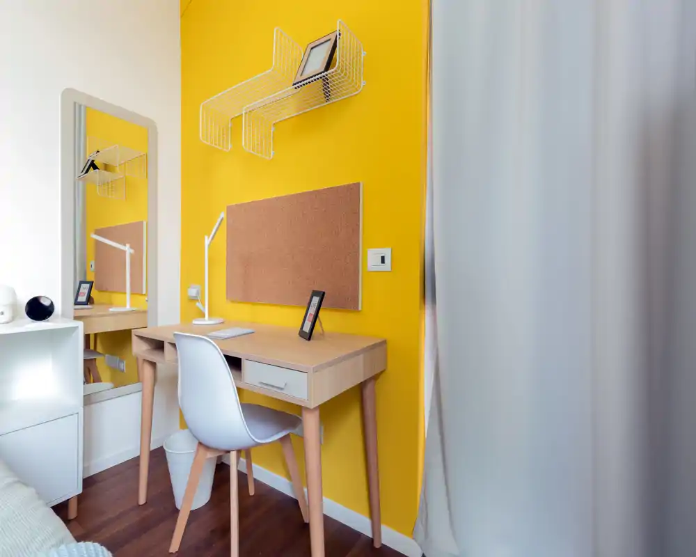 Coin de chambre compact avec un petit bureau en bois devant un mur jaune vif ; chaise, lampe, tableau en liège et étagères—lumineux, moderne et bien rangé, idéal pour étudier ou travailler.