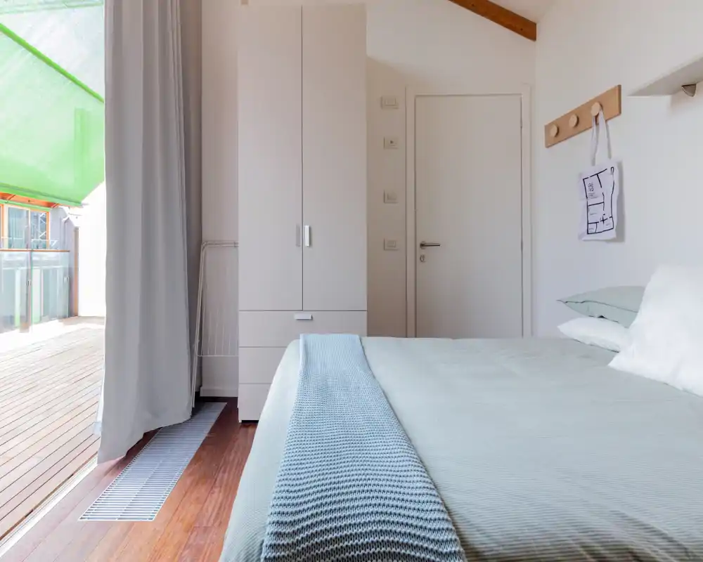 Chambre lumineuse et minimaliste avec lit double bien fait, plaid bleu, armoire et accès direct à un balcon/terrasse ensoleillé.
