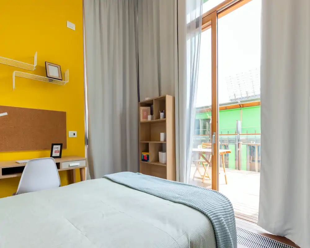 Chambre moderne et lumineuse avec lit bien fait, plaid doux et grandes portes vitrées s'ouvrant sur un balcon — parfaite pour la lumière naturelle.