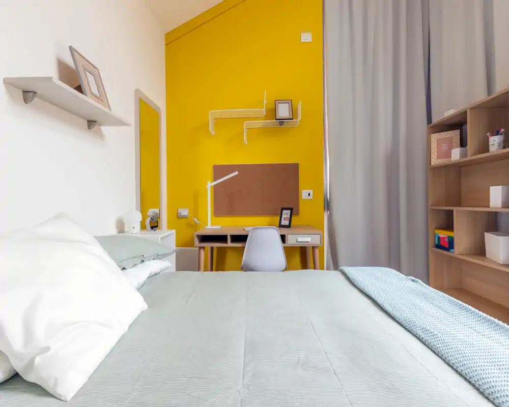 Chambre moderne et lumineuse avec un grand lit au premier plan et un petit bureau contre un mur jaune vif. Lignes épurées, linge de lit doux et étagères organisées créent un espace accueillant et bien mis en scène, parfait pour une annonce.