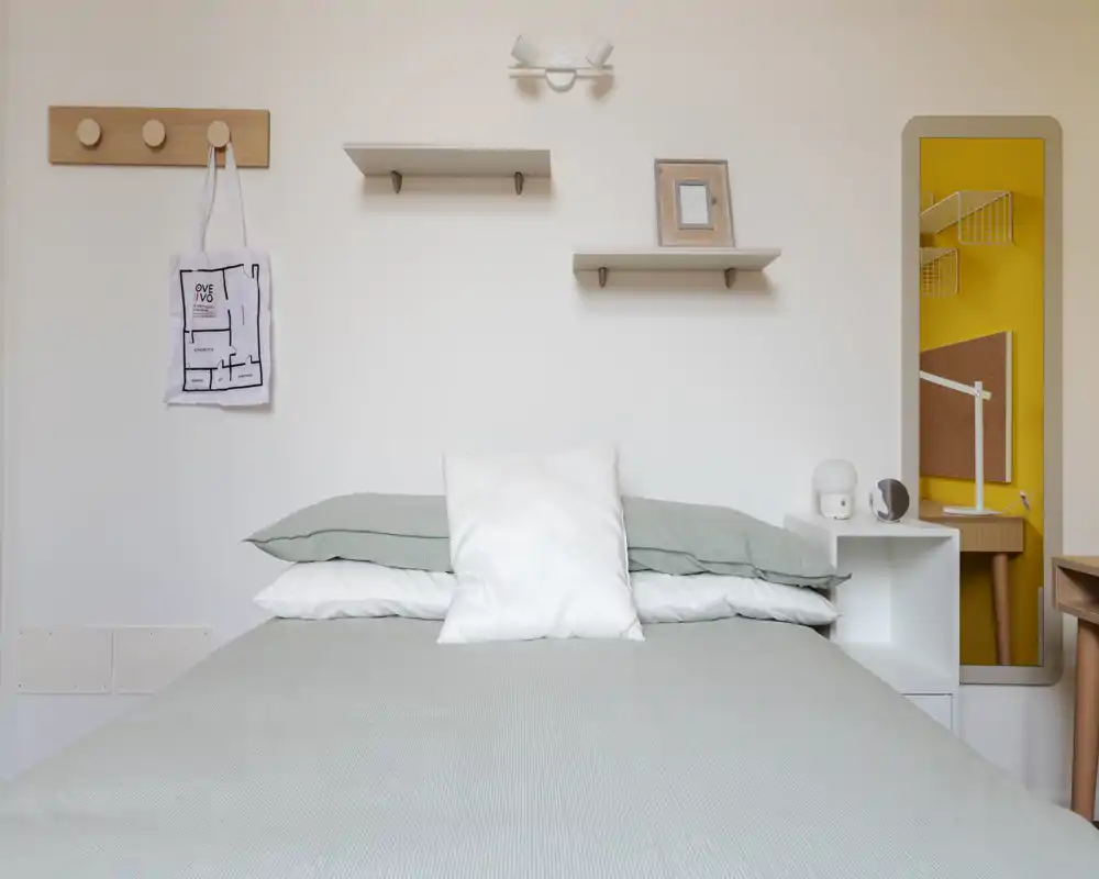 Chambre minimaliste et lumineuse avec lit double centré, linge de lit vert-gris doux, coussin blanc et étagères murales simples ; style épuré et aéré, parfait pour une annonce.