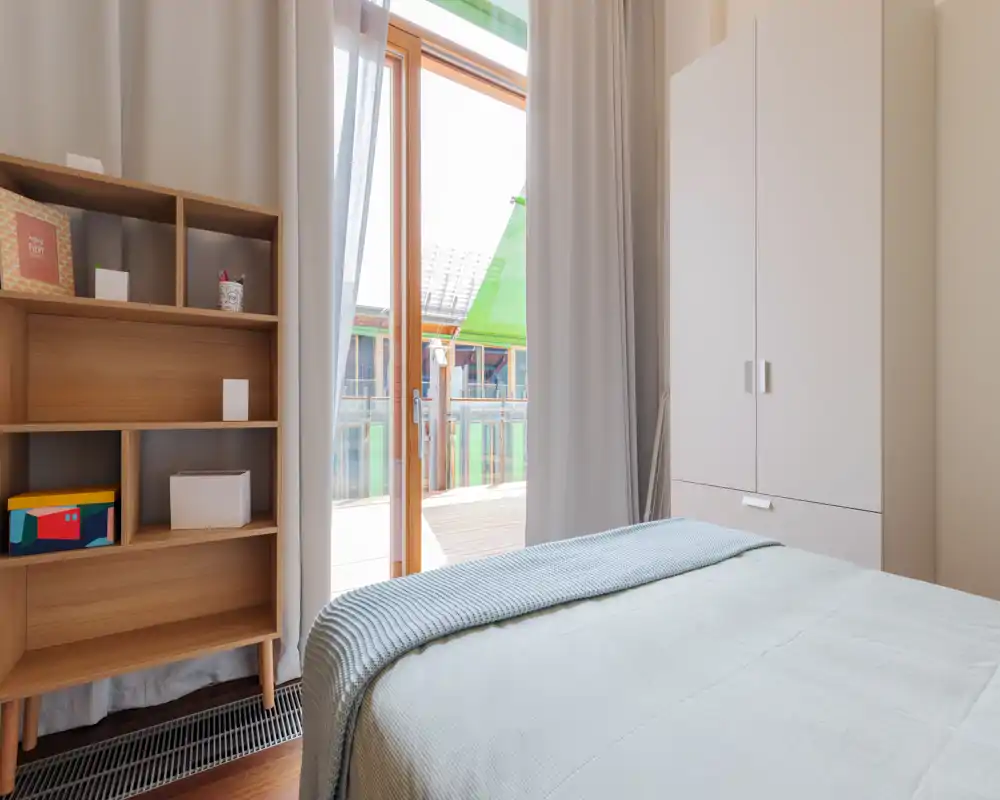 Chambre moderne et lumineuse avec lit bien fait, étagères en bois clair et armoire; grande porte vitrée donnant sur un balcon ensoleillé, apportant beaucoup de lumière naturelle.