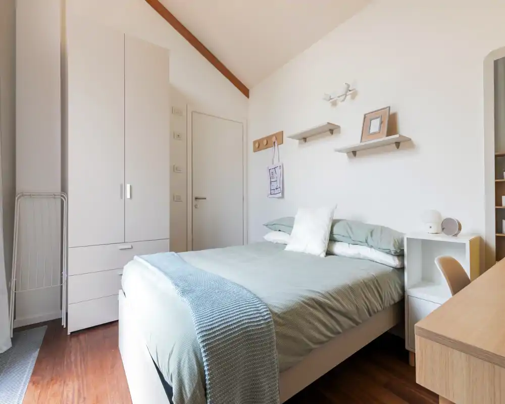 Chambre moderne et lumineuse avec un lit double bien fait, literie vert pâle et étagères minimalistes ; sol en bois chaleureux et petit bureau pour une atmosphère accueillante.