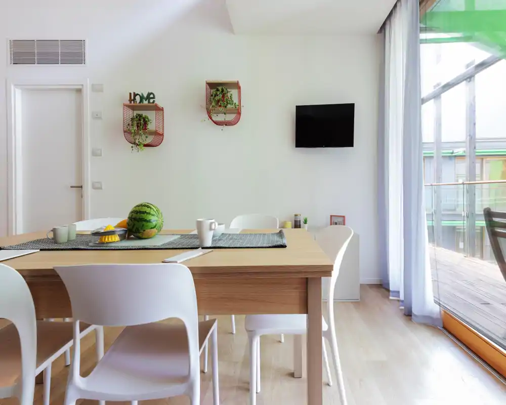 Modern, bright dining area with a wooden table, white chairs and large glass doors providing natural light; simple decorative touches and a clean, minimalist style.
