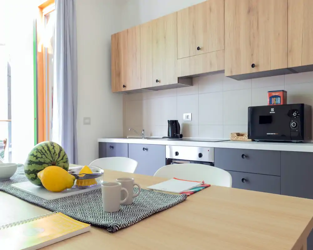 Modern, bright kitchenette with wood upper cabinets, grey lower units, microwave and small appliances. A dining table in the foreground is styled with fruit, mugs and a placemat.