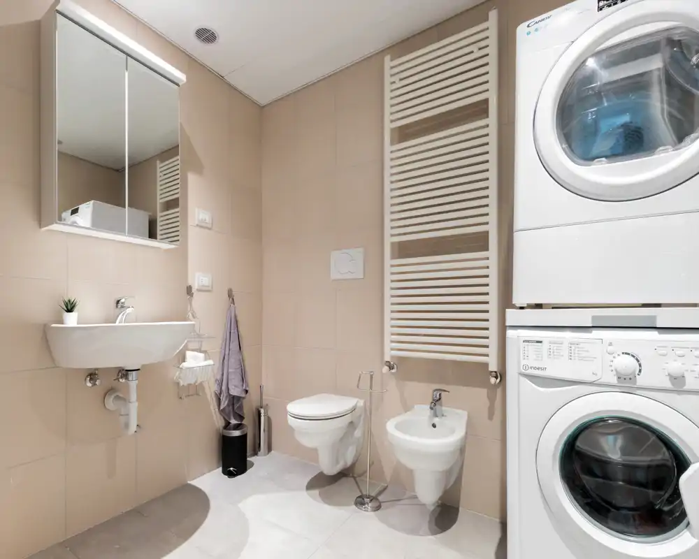 Modern, well-lit bathroom with wall-mounted toilet, bidet, sink with mirror cabinet and stacked washer-dryer units for convenient laundry in the same room.