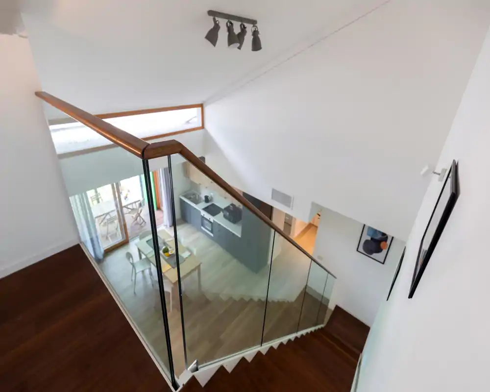 Modern open-plan kitchen seen from a mezzanine: built-in cabinets, stove and a small dining table by sliding glass doors that open onto a balcony. Bright, contemporary space ideal for short-term rentals.
