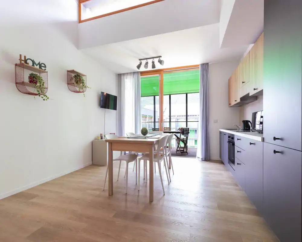 Bright, modern open-plan living area with a dining table and a compact kitchen. Large windows and light wood floors create an airy, welcoming space ideal for city stays.