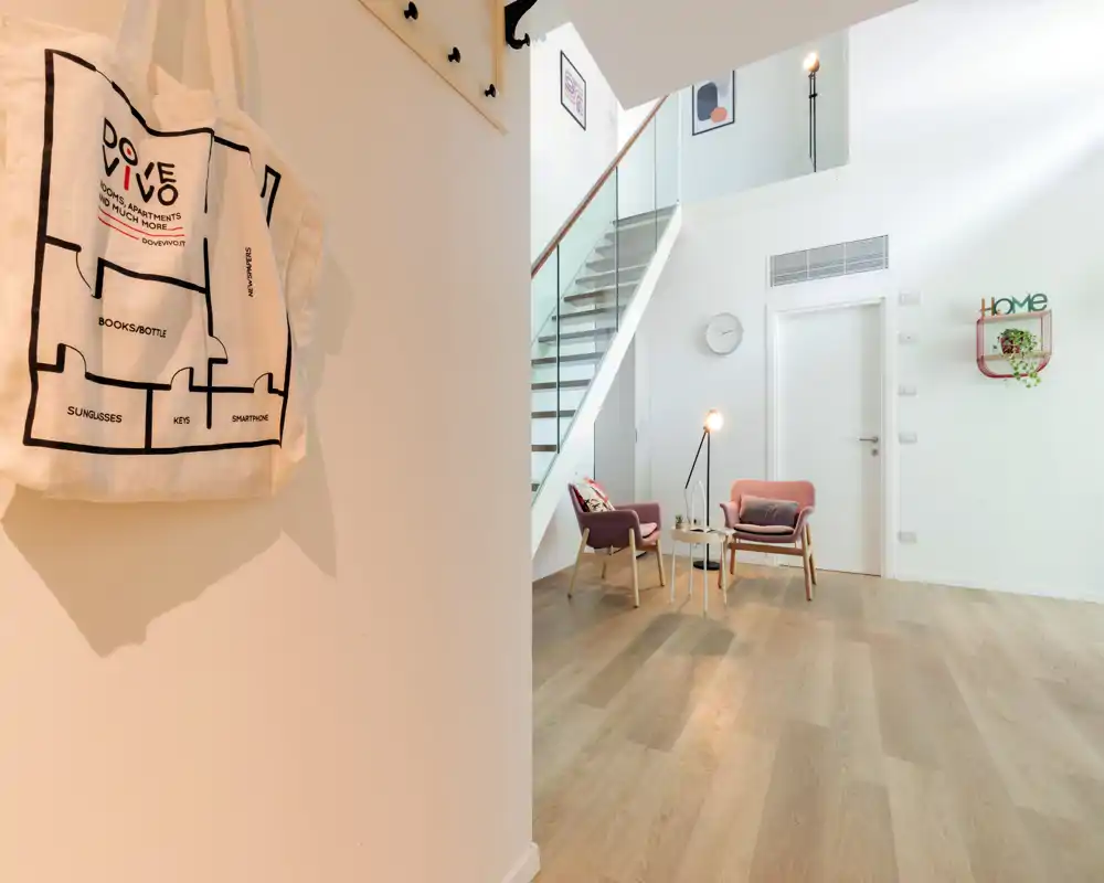 Bright, modern entrance area with light oak flooring, two accent chairs beside a small table and a staircase leading to an upper level. Minimalist decor and soft natural light create a welcoming arrival space.