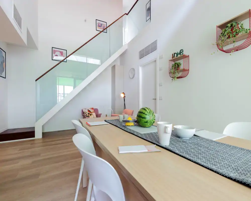 Modern dining area with a long wooden table set with a runner, bowls and a decorative watermelon; bright, airy space with contemporary chairs and plants on the wall.