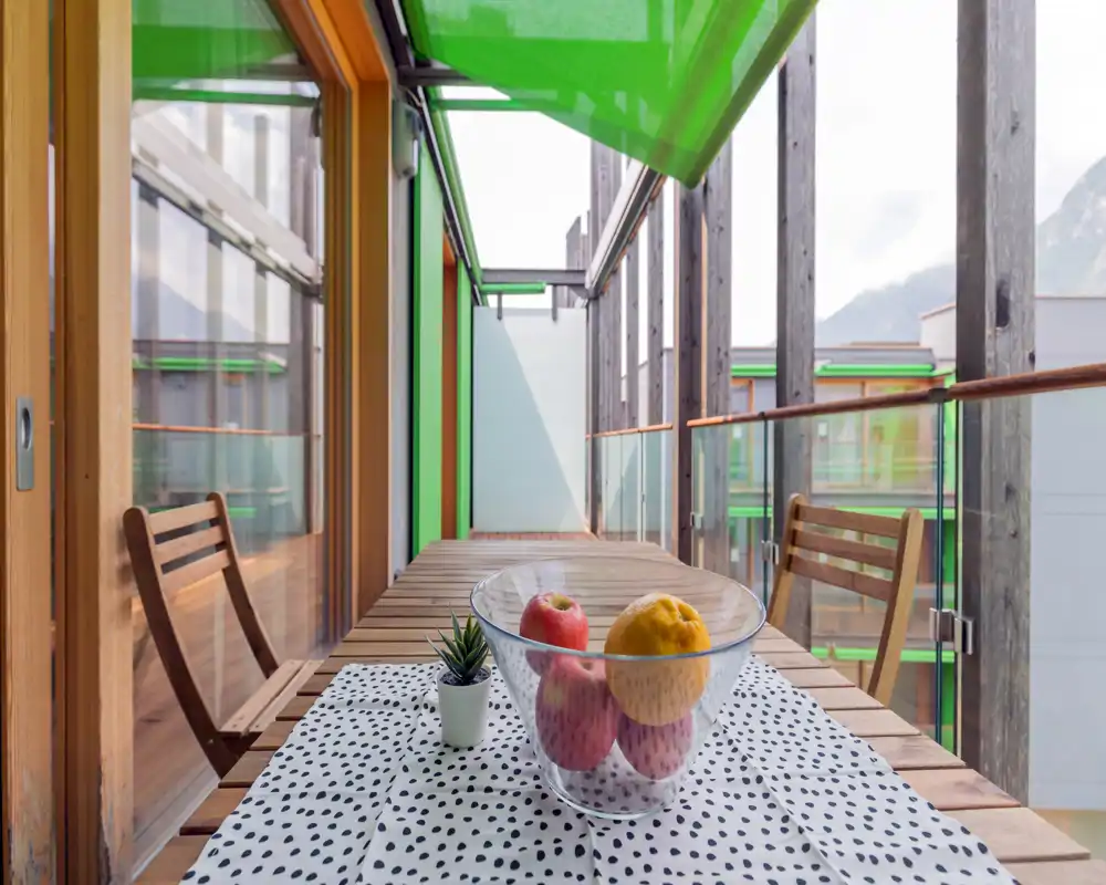 Sunny balcony with a wooden table and chairs, glass railing and a decorative bowl of fruit — ideal for morning coffee with mountain views.