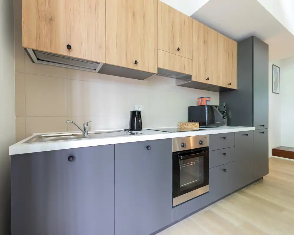 Modern fitted kitchen with gray lower cabinets and light wood upper cabinets, built-in oven, sink and countertop appliances; clean, bright and functional space ideal for short-term rentals.