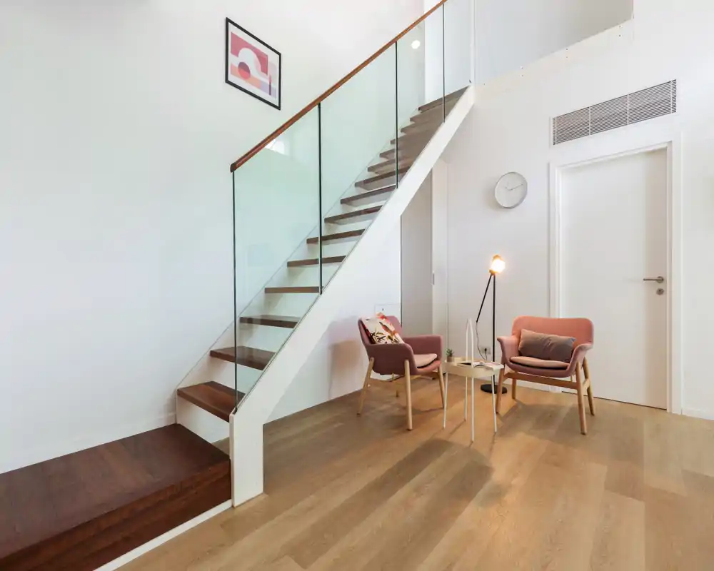 Bright, modern living area under a staircase featuring two cozy armchairs, a small coffee table and a floor lamp — ideal for a reading nook or conversation corner.