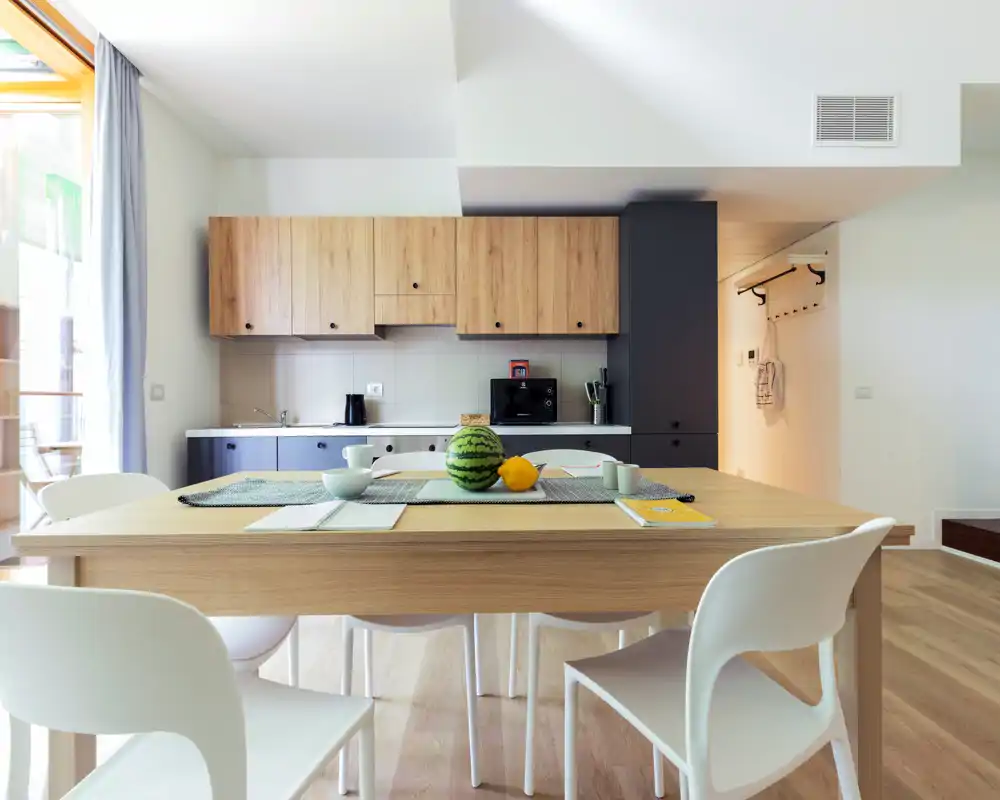 Modern open kitchen with wooden cabinets, a central dining table set with simple tableware and fruit; bright natural light and minimalist styling create a welcoming, functional space.