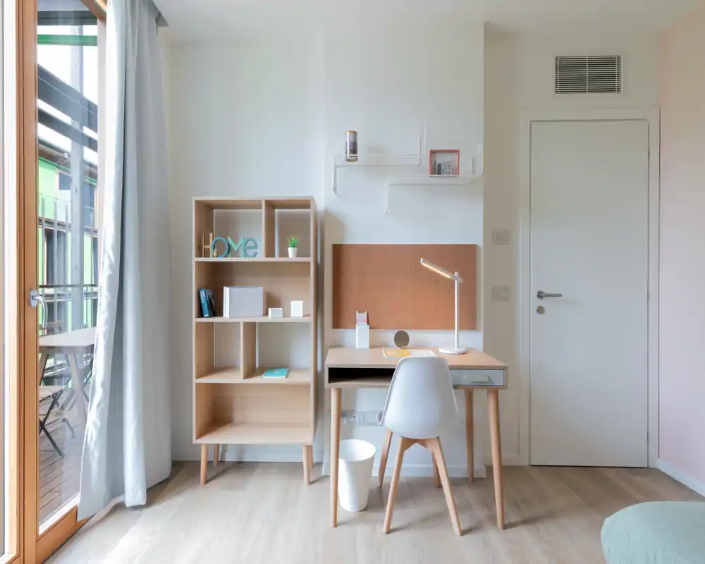 Bright, minimal bedroom workspace with a wooden desk, chair and shelving next to a balcony door — ideal for remote work in a cozy apartment.