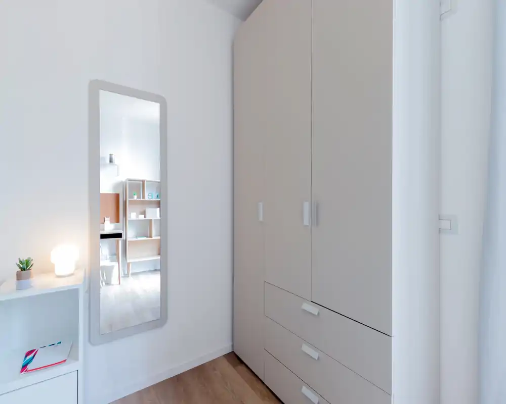 Compact bedroom corner with a tall wardrobe, wall mirror and bedside shelf; bright, minimal and neatly styled.