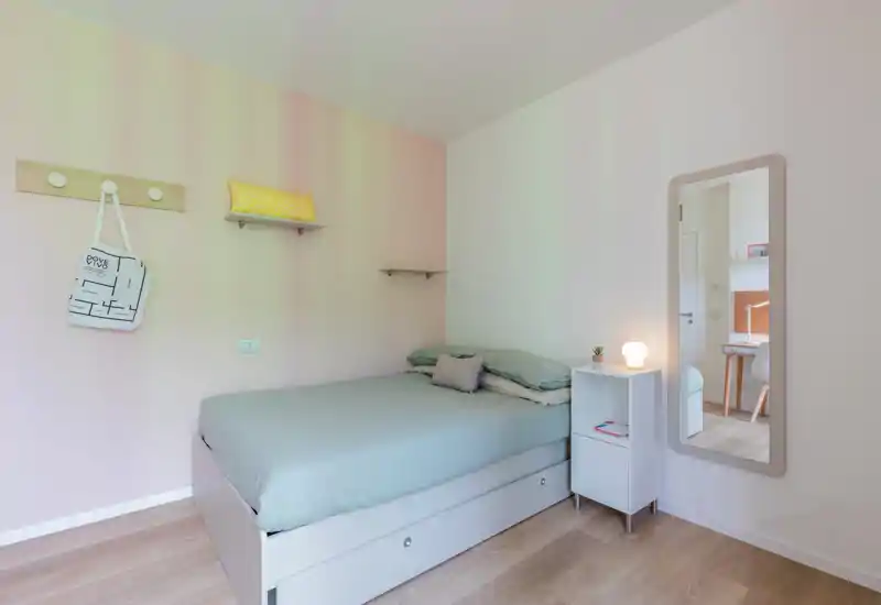 Dormitorio luminoso y ordenado con cama verde claro, estante de noche y espejo de cuerpo entero; paredes en tonos pastel y suelo de madera ofrecen un ambiente minimalista y acogedor, perfecto para un anuncio de alquiler.