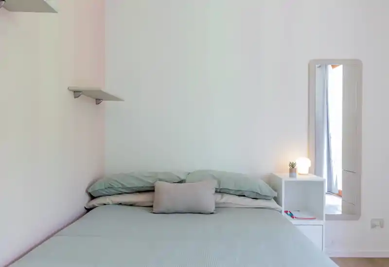 Dormitorio luminoso y minimalista con cama doble bien hecha, ropa de cama en tonos pastel, estantería de noche y un espejo de cuerpo entero que refleja la luz natural.