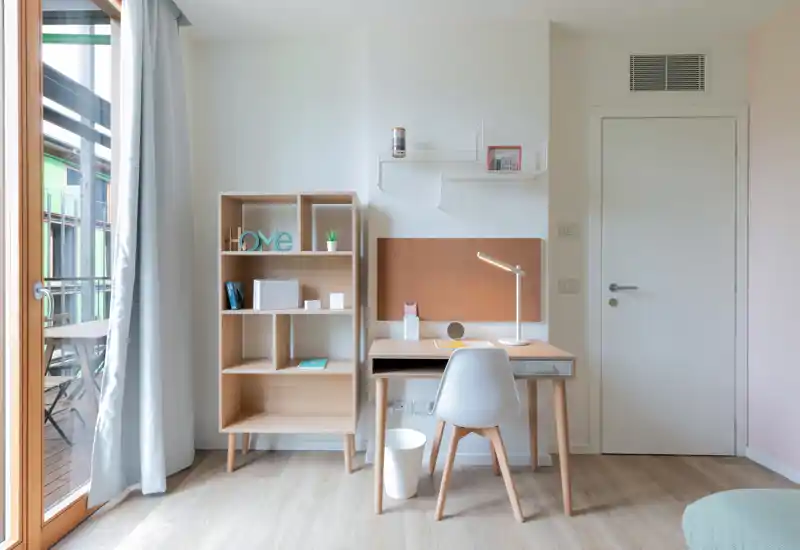 Espacio de trabajo luminoso y minimalista en la habitación con escritorio de madera, silla y estantería junto a la puerta del balcón — ideal para teletrabajo.