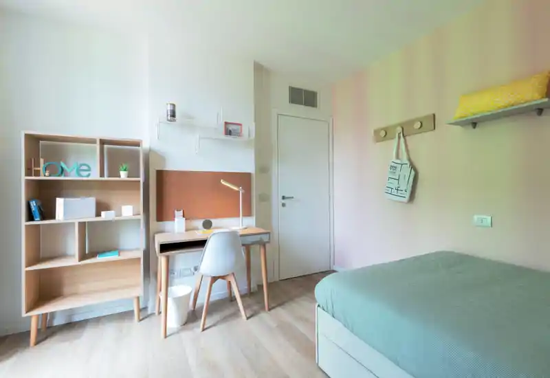 Habitación individual luminosa y ordenada con cama a la derecha, escritorio de madera y estantería abierta — ideal para estudiante o ocupante individual.