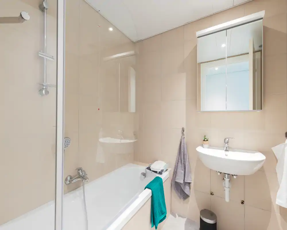 Bright, modern bathroom with a bathtub/shower combo, wall-mounted sink, mirrored cabinet and neutral beige tiles.