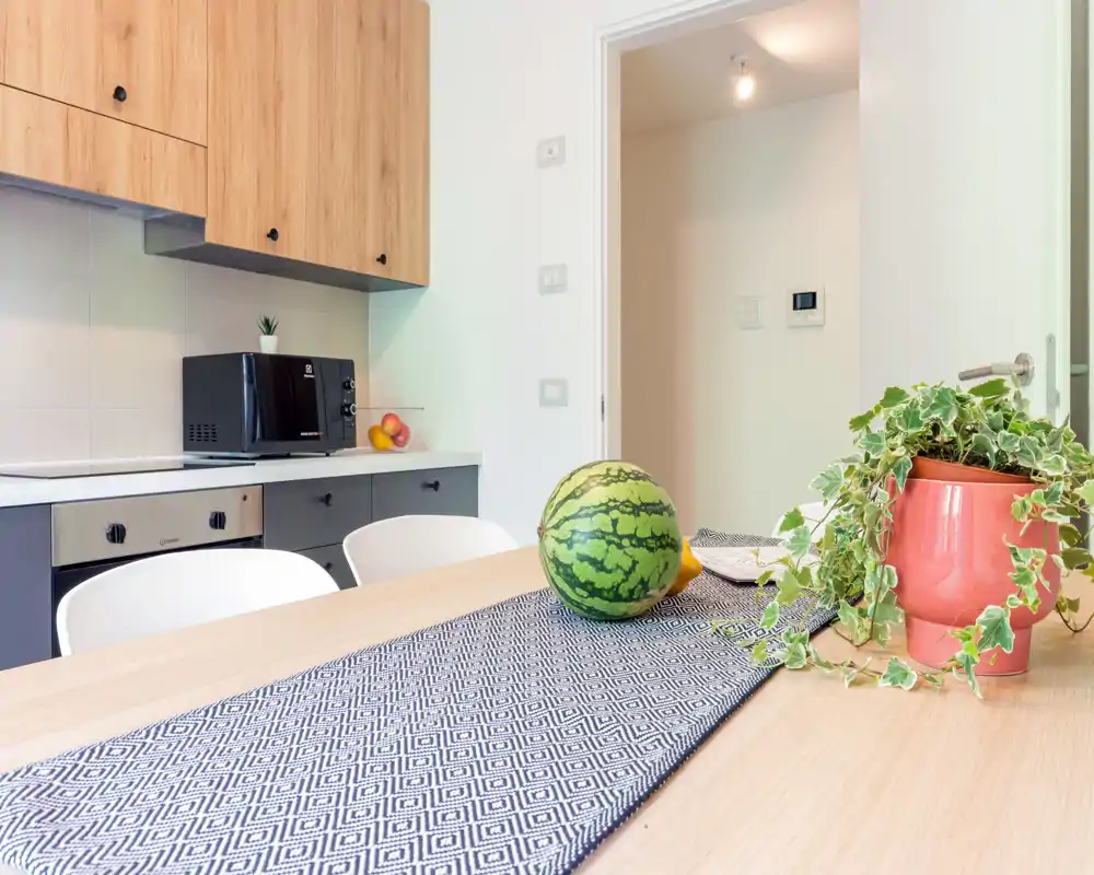 Modern kitchen corner with a wooden dining table in the foreground, decorative runner, a potted plant and fresh fruit; built-in oven and microwave visible along the worktop.