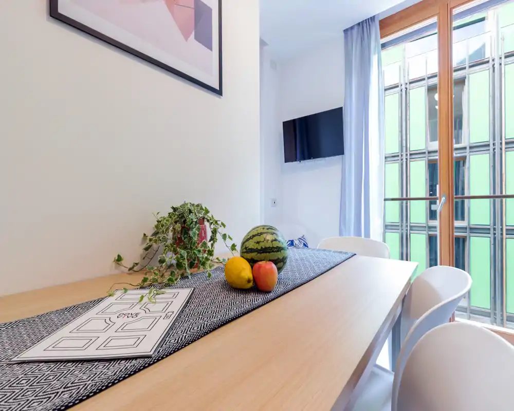 Bright, modern dining area with a wooden table, white chairs, decorative runner, potted plant and fresh fruit, next to a large window that fills the space with natural light.