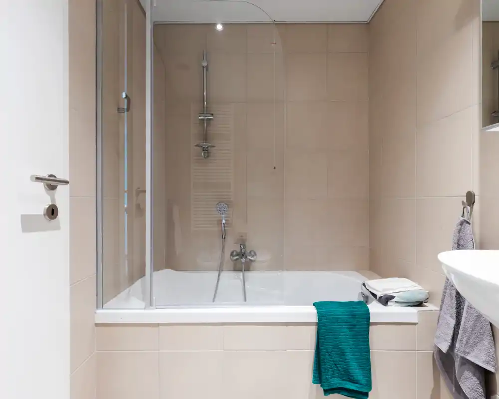 Modern bathroom with a tiled bathtub and glass shower screen, chrome shower fixtures and a clean white sink visible at the right edge.