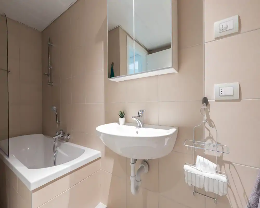 Clean, modern bathroom with a bathtub, wall-mounted sink and mirrored cabinet; neutral beige tiles and organized accessories provide a bright, uncluttered feel.