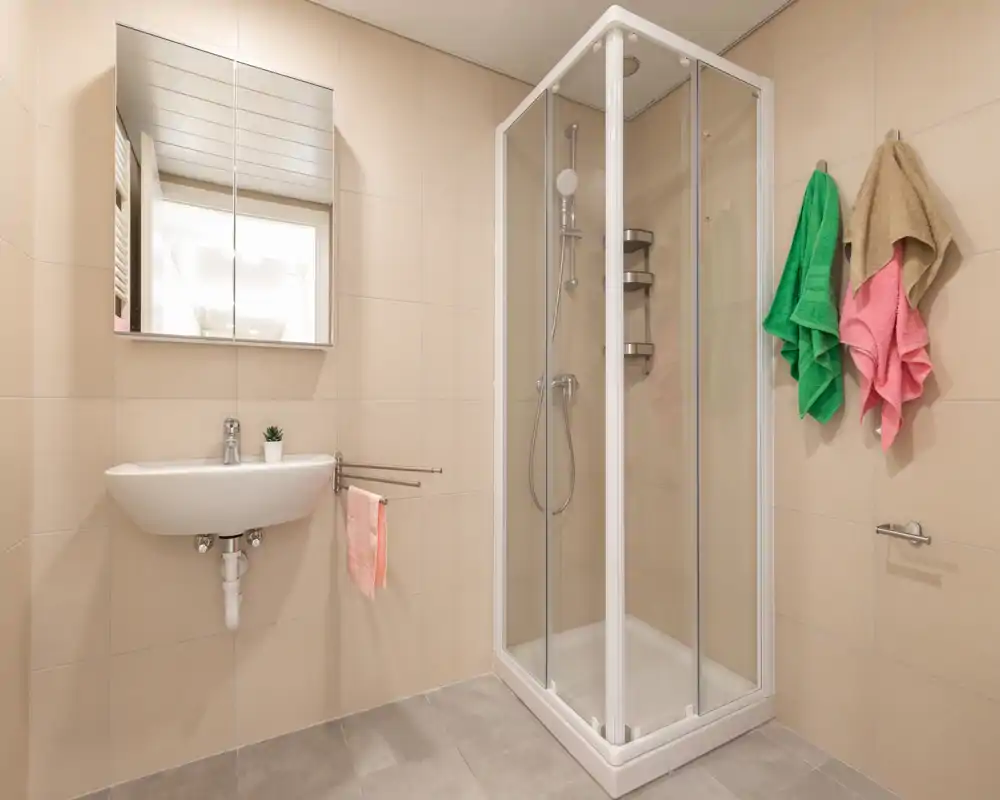 Small modern bathroom with a corner glass shower, wall-mounted sink and mirrored cabinet. Neutral tiles and colorful towels add a fresh, tidy look.