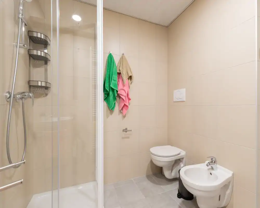 Bright, modern bathroom with shower cabin, wall-mounted toilet and bidet; neutral tiles and hanging towels create a clean, functional space.