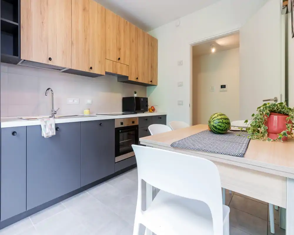 Modern fitted kitchen with wooden upper cabinets and matte lower units, integrated oven and a dining table set with plants and fruit—bright, tidy and ready for use.