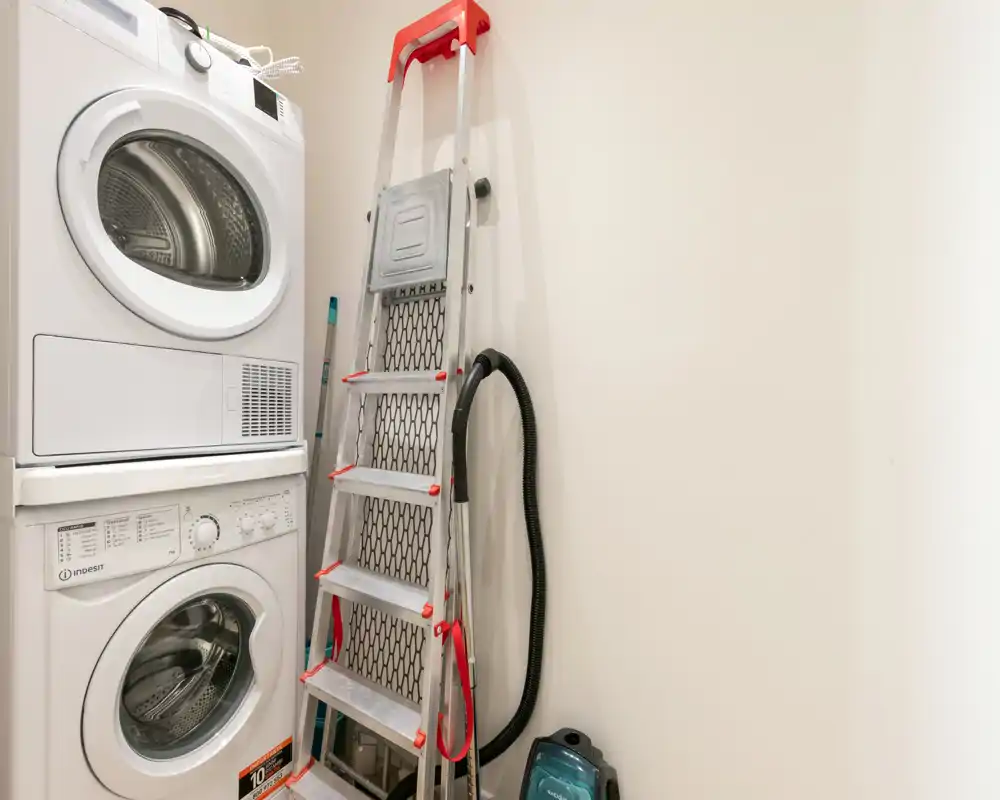 Compact laundry area with stacked washer and dryer; tidy utility corner including a ladder and vacuum cleaner. Bright neutral walls and efficient vertical appliance layout make good use of space.