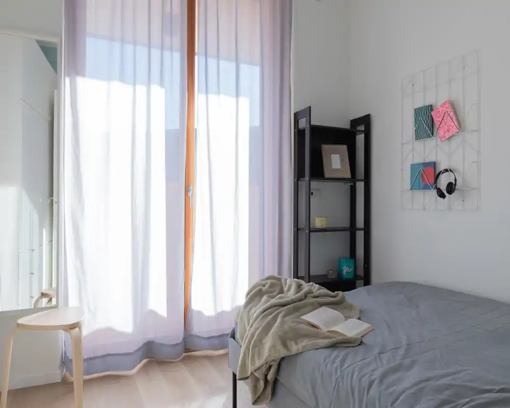 Chambre lumineuse et minimaliste avec lit soigné, plaid doux et livre ouvert près d'une grande fenêtre ensoleillée — idéale pour un séjour confortable en ville.