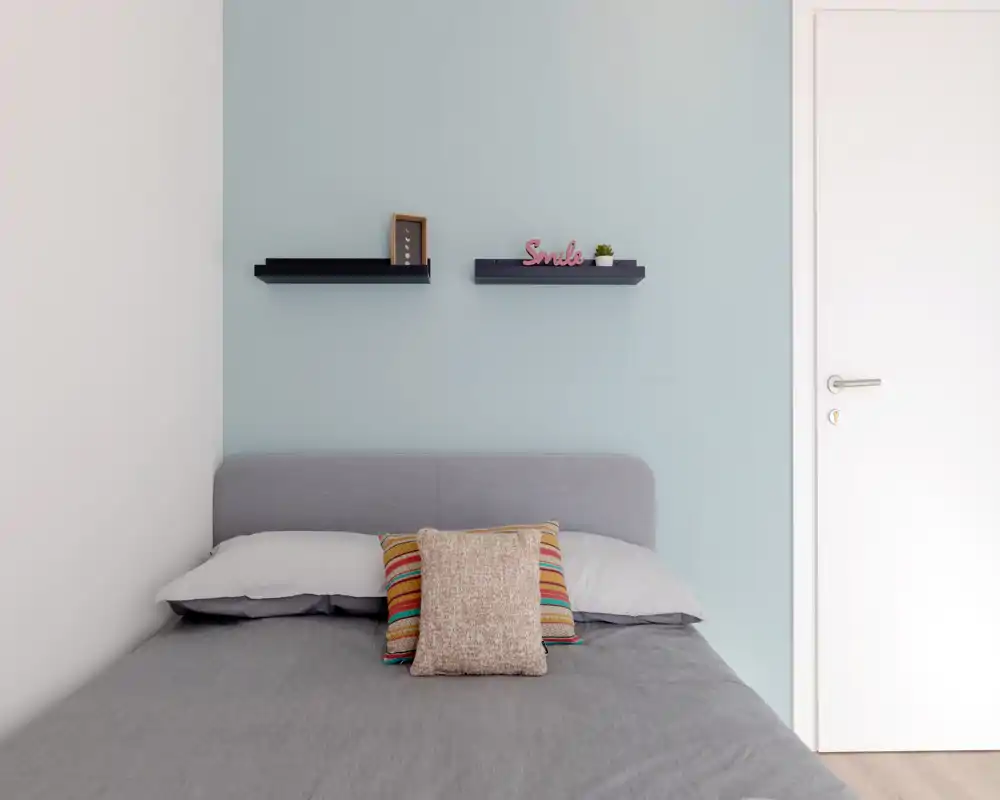 Chambre lumineuse et minimaliste avec un lit double bien fait, linge de lit gris et coussins colorés sur un mur bleu pâle agrémenté de petites étagères flottantes.