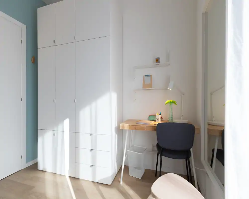 Coin de chambre lumineux avec une grande armoire blanche, un petit bureau en bois et une chaise, un miroir et une lumière naturelle douce — idéal pour une personne seule ou un coin bureau.