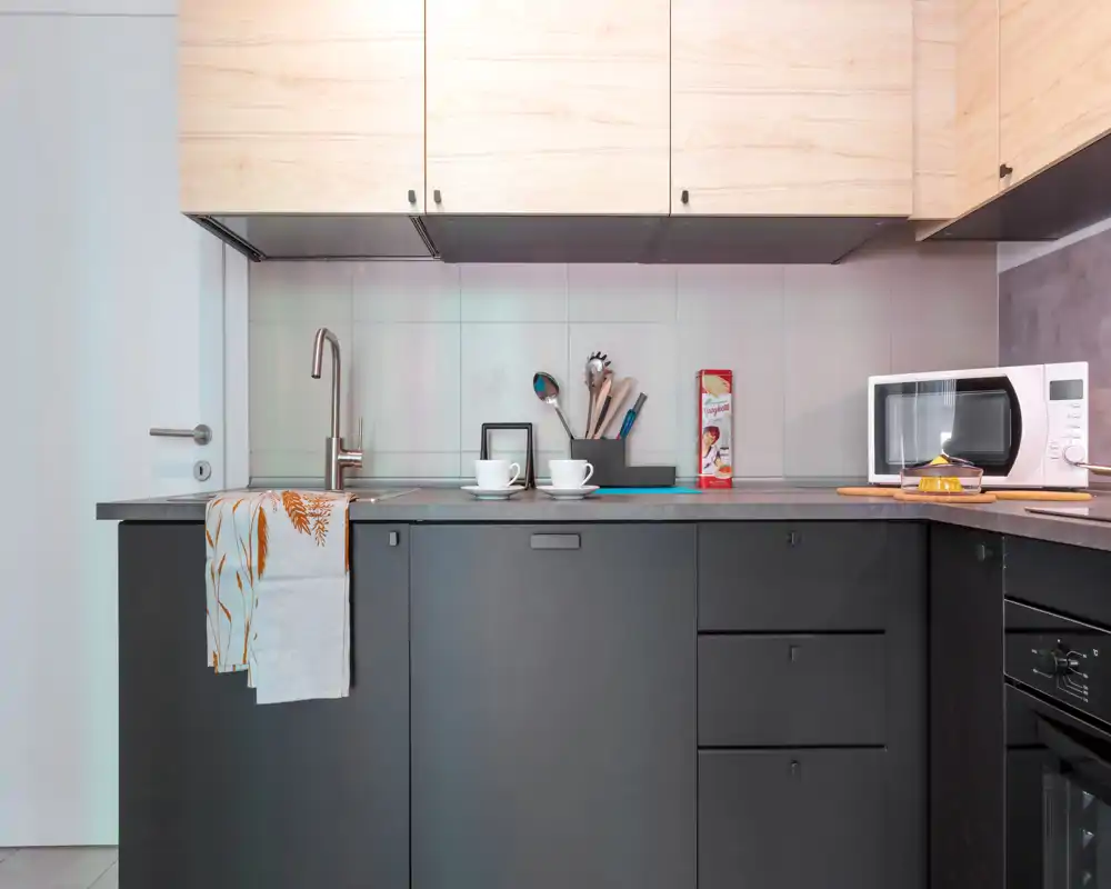 Cuisine compacte moderne avec meubles bas mats foncés, placards hauts en bois clair, évier encastré et petits appareils sur le plan de travail. Mise en scène soignée avec tasses, ustensiles et torchon pour une ambiance accueillante et fonctionnelle.