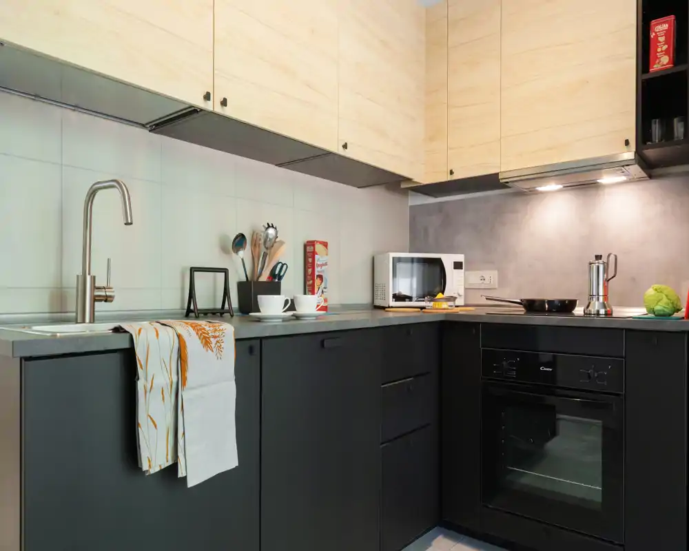 Cuisine compacte moderne avec meubles bi‑ton, four encastré, micro‑ondes et plan de travail avec évier et ustensiles — bien éclairée et soignée, idéale pour cuisiner confortablement.
