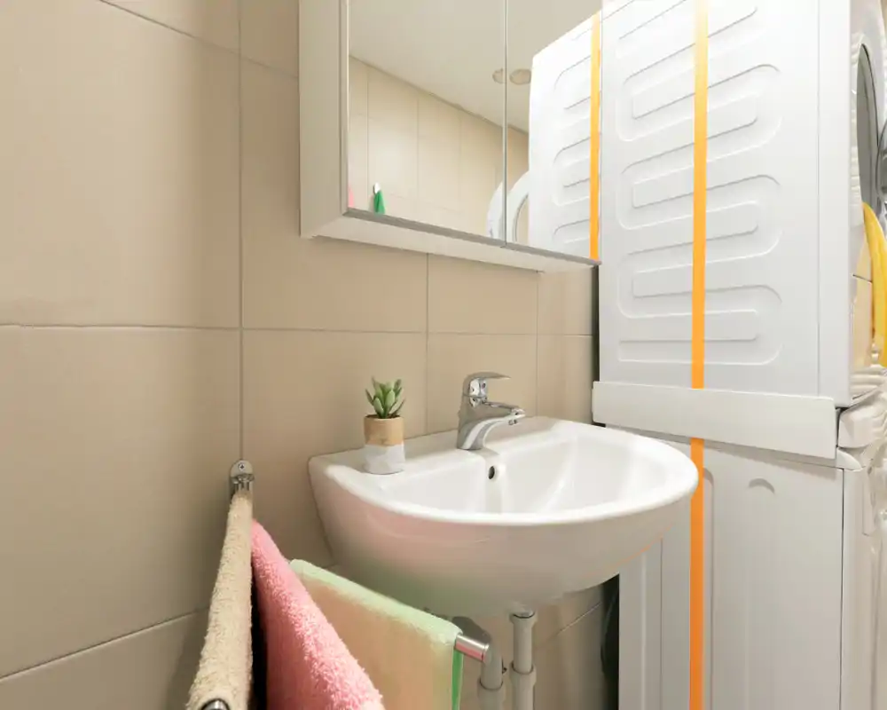 Salle de bains compacte et propre avec lavabo mural, miroir armoire et lave-linge/sèche-linge empilés. Carrelage neutre et éclairage doux pour un espace fonctionnel et soigné.