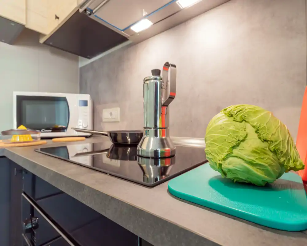Plan de travail de cuisine moderne avec plaque à induction, cafetière moka, poêle, micro-ondes et chou sur une planche — propre, bien éclairé et prêt à cuisiner.