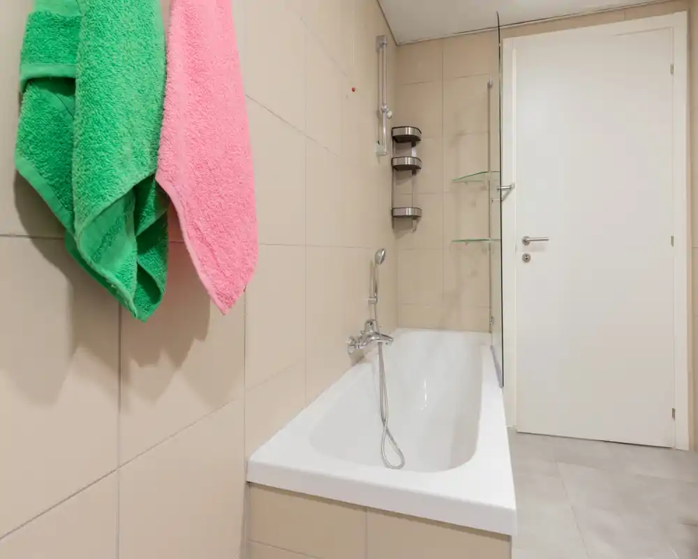 Salle de bains moderne et propre avec baignoire, douchette à main et carrelage beige clair ; lumineuse et bien rangée.