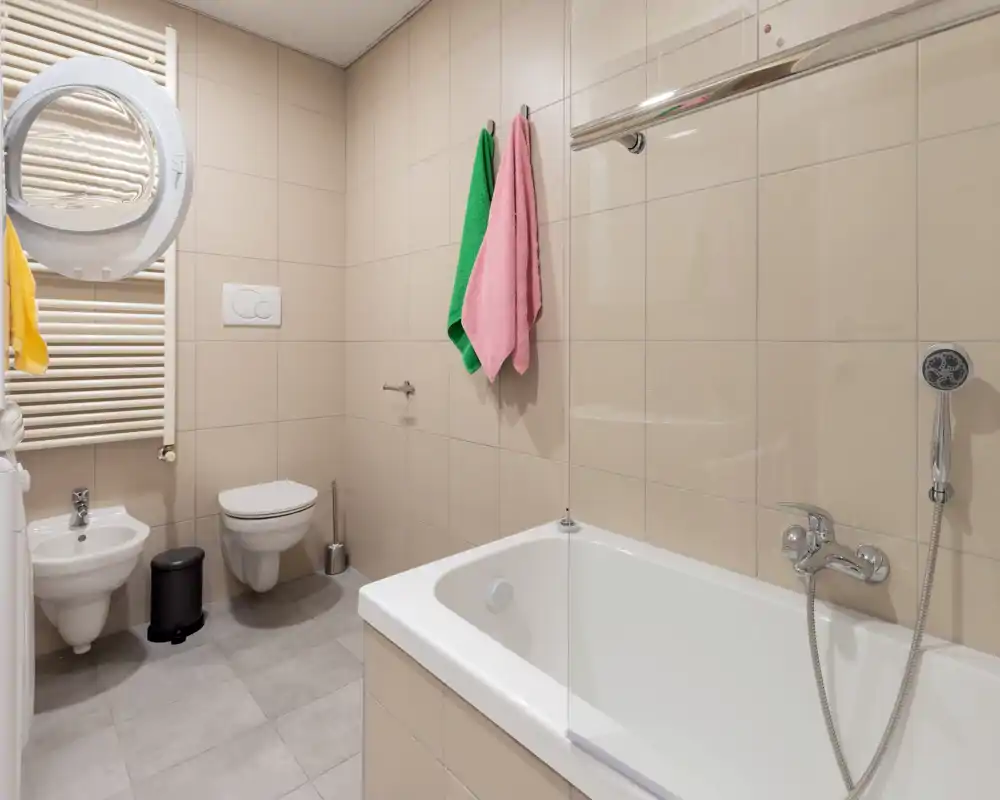 Salle de bains moderne et lumineuse avec baignoire, douchette, WC suspendu et bidet, porte-serviettes et carreaux beiges aux finitions nettes.