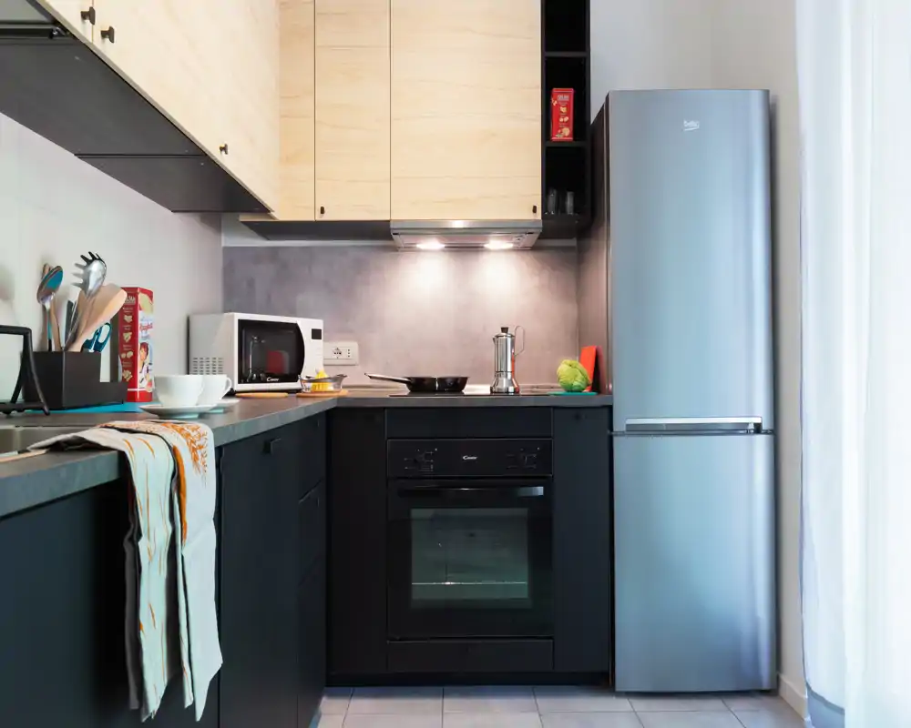Cuisine compacte et moderne avec éléments bas noirs, placards hauts en bois clair, four, micro-ondes et réfrigérateur en inox — propre, lumineuse et fonctionnelle.